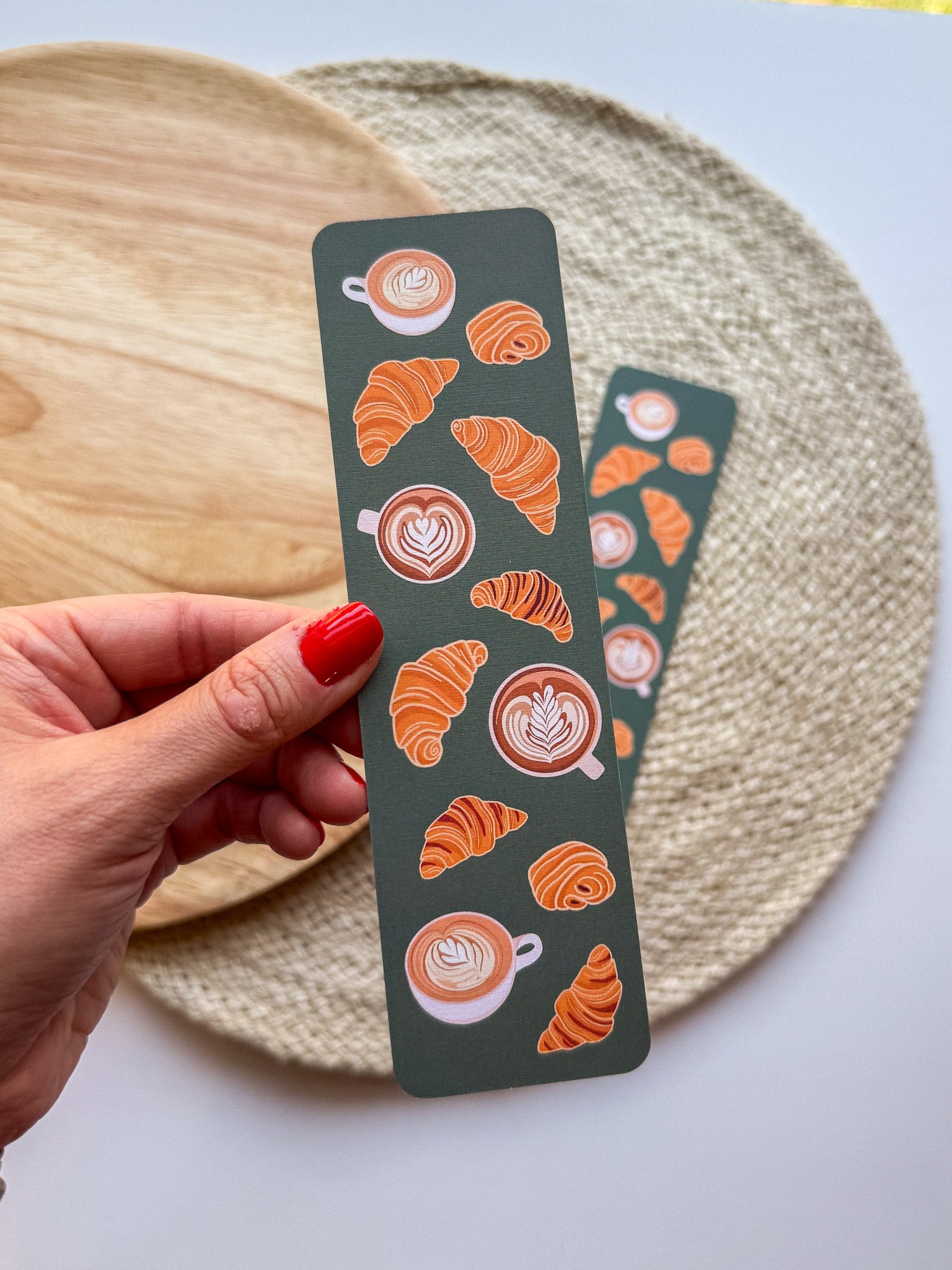 Hand holding a decorative bookmark with coffee and croissant pattern designs on a light background