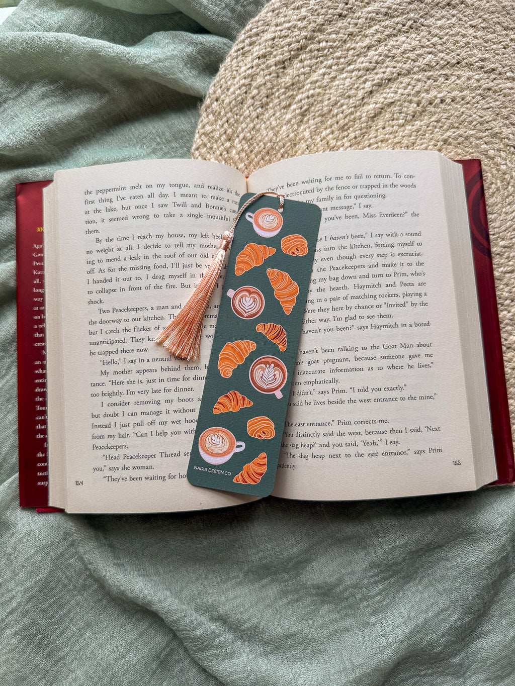 Open book with a decorative bookmark featuring croissant and latte coffee illustrations and a tassel on a textured surface.