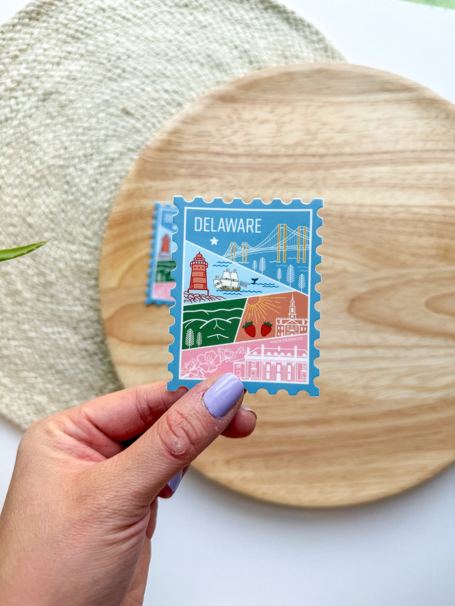 illustrated Delaware-themed stamp shaped sticker held by a hand on a wooden surface