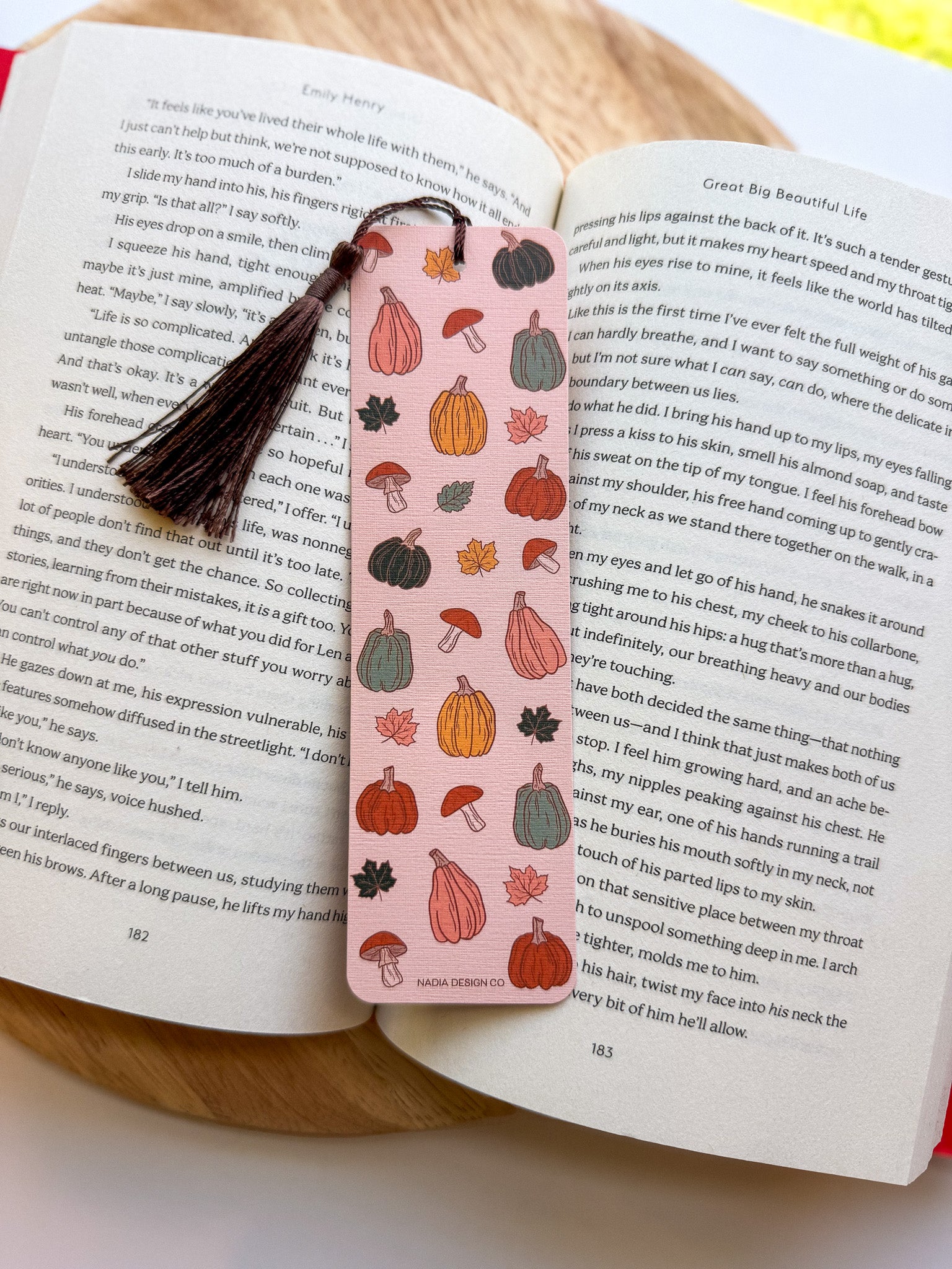 Open book with two bookmarks featuring autumn-themed illustrations with brown tassel on a woven surface.