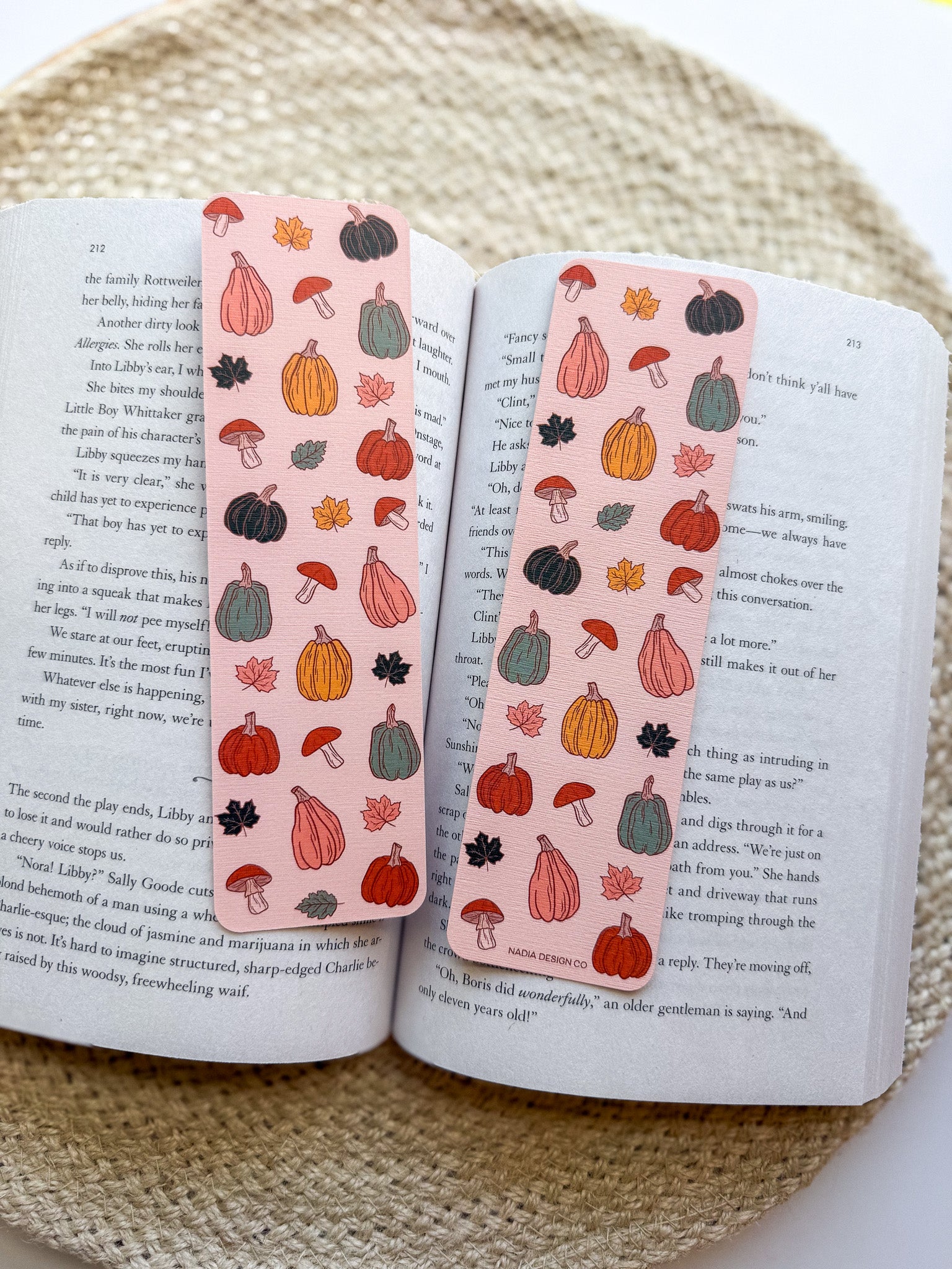 Open book with two bookmarks featuring autumn-themed illustrations on a woven surface.