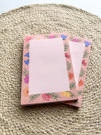 A stack of pink notepads with multicolored floral patterns on the edges, placed on a beige textured surface.
