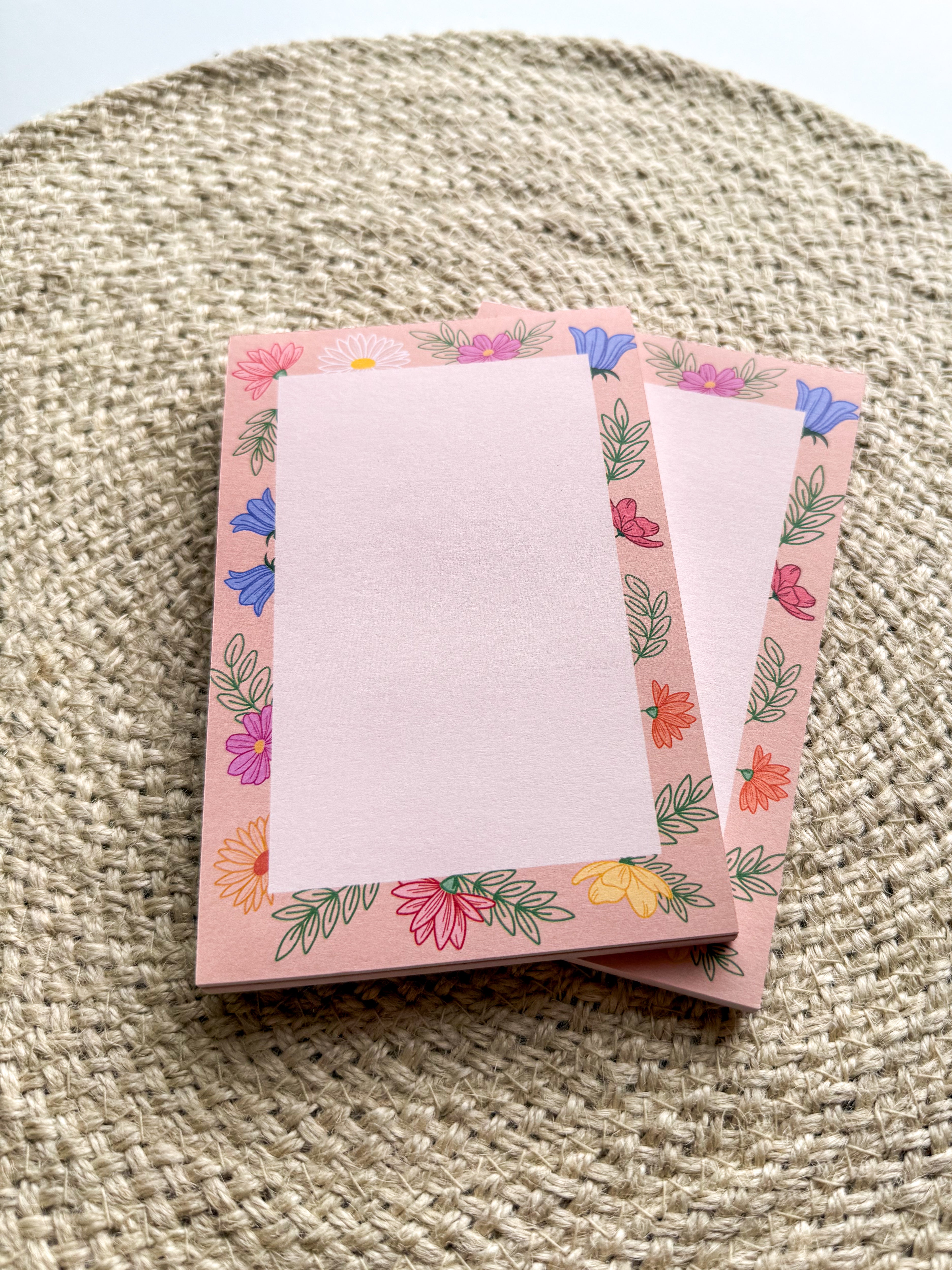 A stack of pink notepads with multicolored floral patterns on the edges, placed on a beige textured surface.