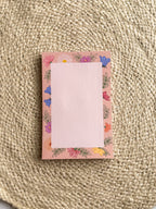 A stack of pink notepads with multicolored floral patterns on the edges, placed on a beige textured surface.
