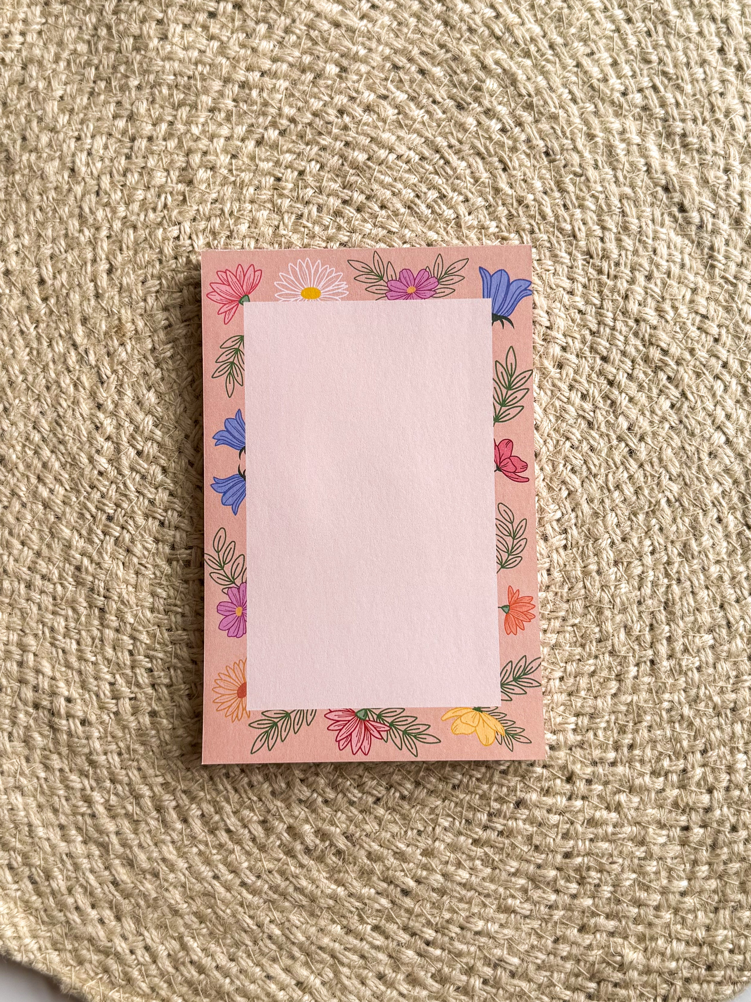 A stack of pink notepads with multicolored floral patterns on the edges, placed on a beige textured surface.