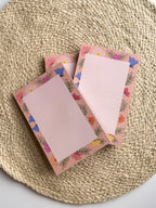 A stack of pink notepads with multicolored floral patterns on the edges, placed on a beige textured surface.