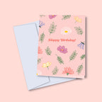 A greeting card with a floral birthday bouquet illustration and the text 'Happy Birthday!' on the front, accompanied by a white envelope, against a pink background.