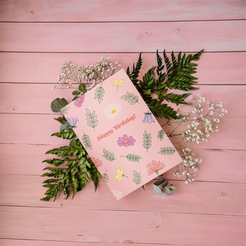 A greeting card with a floral birthday bouquet illustration and the text 'Happy Birthday!' on the front, accompanied by a white envelope, against a pink background.
