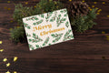 A Christmas card with 'Merry Christmas' text and a green leaf pattern, placed on a wooden surface with pine cones and golden stars around it.