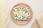 A Christmas card with 'Merry Christmas' text and a green leaf pattern, placed on a wooden surface with pine cones and golden stars around it.