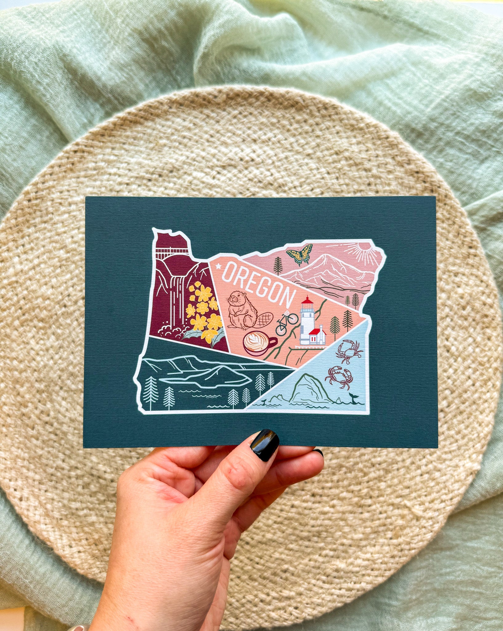 Postcard with a colorful illustration of Oregon held by a hand on a textured surface.