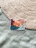 Colorful illustrated sticker shaped like the state of Oregon on a textured surface