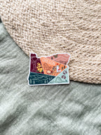 Colorful illustrated sticker shaped like the state of Oregon on a textured surface