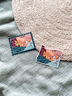 Colorful illustrated stickers. one shaped like the state of Oregon and the other stamp shaped on a textured surface 