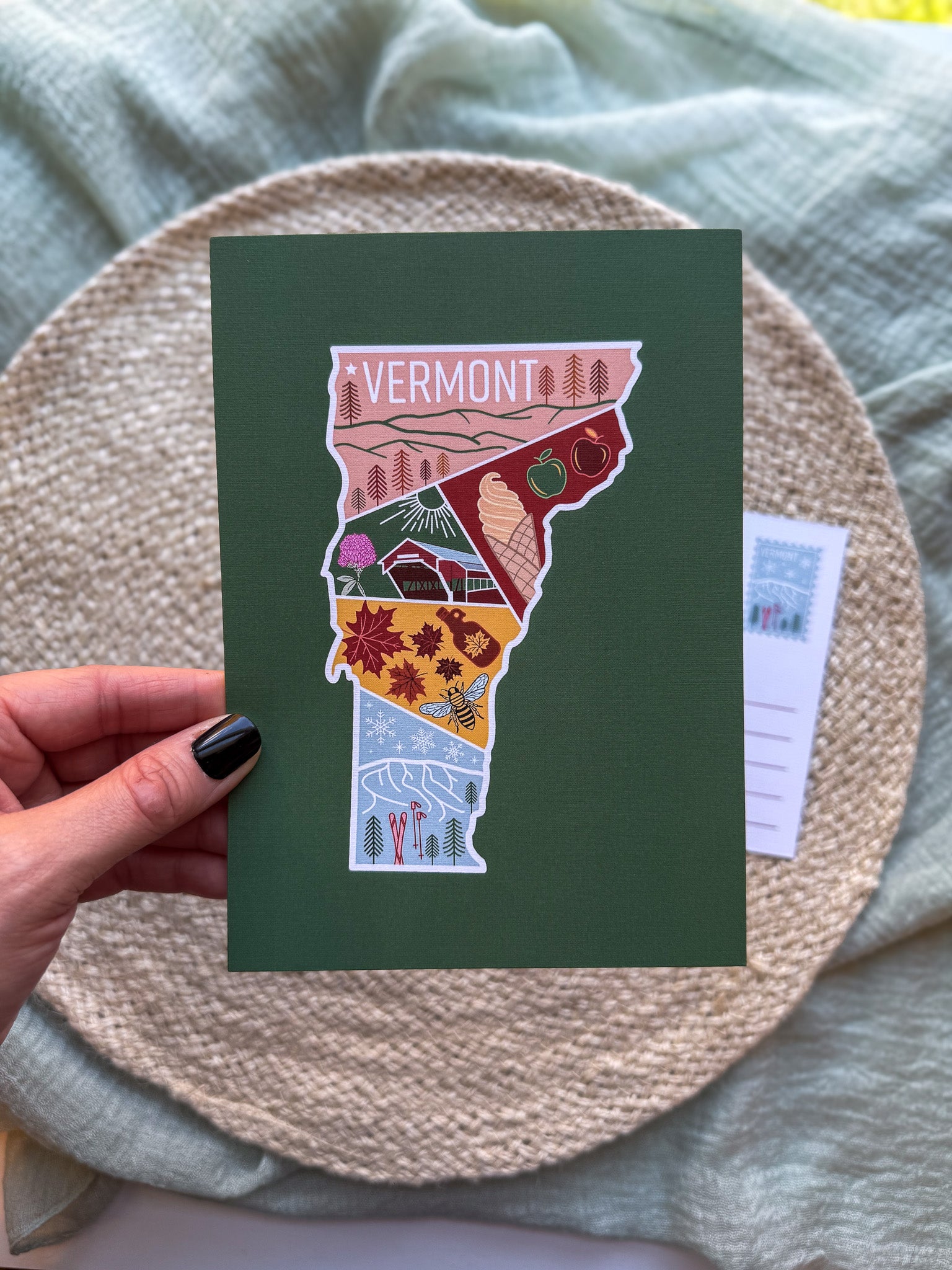 Postcard with a colorful illustration of Vermont held by a hand on a textured surface.