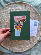 Postcard with a colorful illustration of Vermont held by a hand on a textured surface.