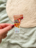 Hand holding a colorful Vermont-shaped sticker against a textured beige background.