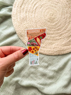 Hand holding a colorful Vermont-shaped sticker against a textured beige background.