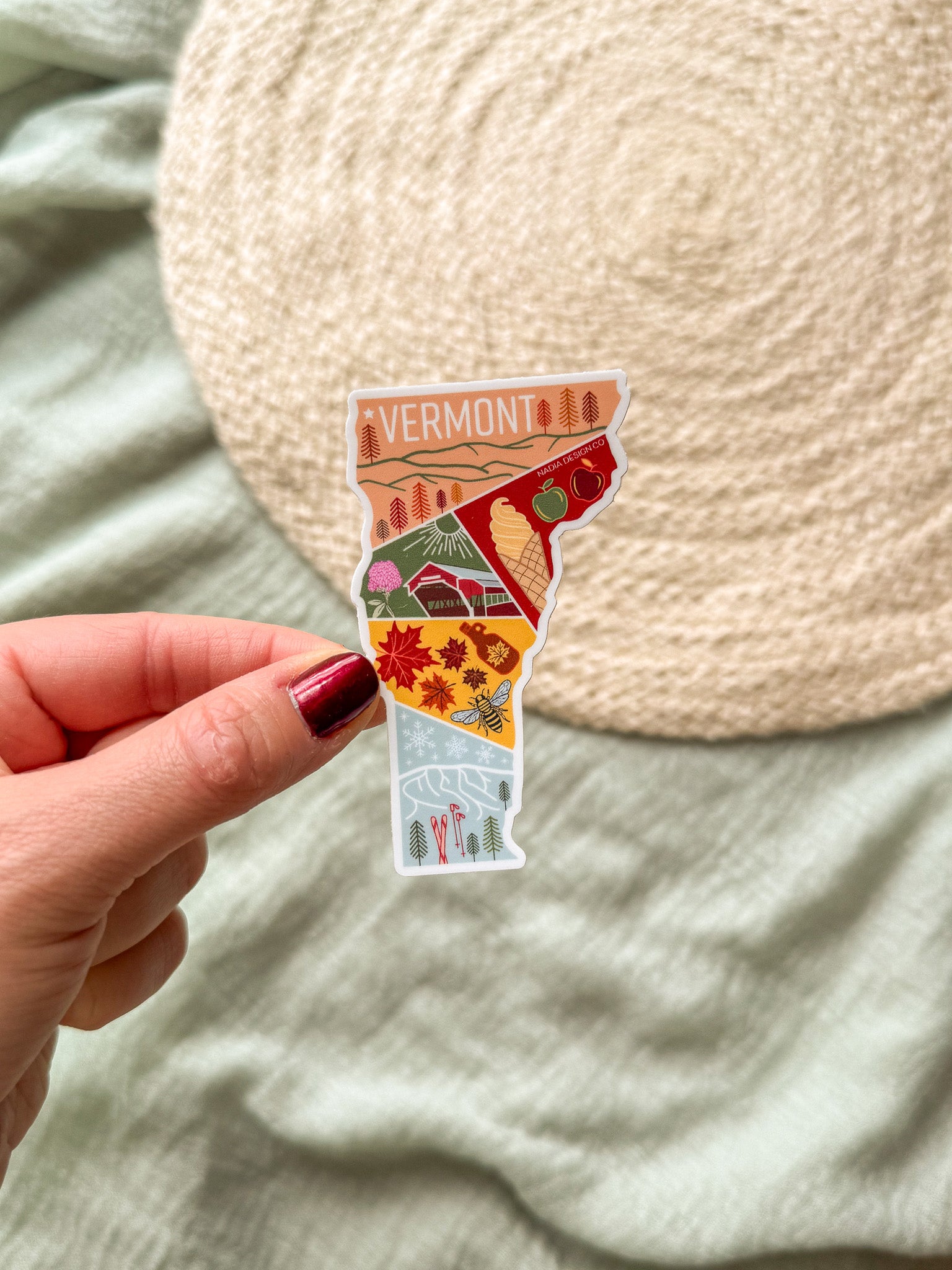 Hand holding a colorful Vermont-shaped sticker against a textured beige background.