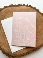 A pink greeting card with the text 'I LOVE YOU MOM' and a floral design, specifically tulips, lying on a white surface with a white envelope beside it and on a wood background.
