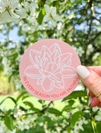 A pink vinyl sticker with a floral design and the text 'In every season you are faithful' held against a backdrop of trees and flowers.