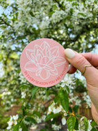 A pink vinyl sticker with a floral design and the text 'In every season you are faithful' held against a backdrop of trees and flowers.
