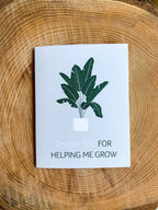 A greeting card with a green and white illustration of a plant, held in a hand with a pink floral background.