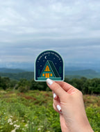 A sticker with an illustration of a cabin under the moon and stars held against a blurred naturxal background.