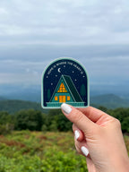 A sticker with an illustration of a cabin under the moon and stars held against a blurred natural background.