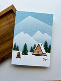 A greeting card depicting a winter scene with a cabin, snow-covered trees, mountains in the background, and a bear in the foreground. The card is placed next to a wooden object, and an envelope with a plastic sleeve is visible.
