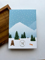 back of a greeting card depicting a winter scene with a cabin, snow-covered trees, mountains in the background, and a bear in the foreground. The card is placed next to a wooden object, and an envelope with a plastic sleeve is visible.
