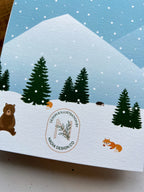 A greeting card depicting a winter scene with a cabin, snow-covered trees, mountains in the background, and a bear in the foreground. The card is placed next to a wooden object, and an envelope with a plastic sleeve is visible.