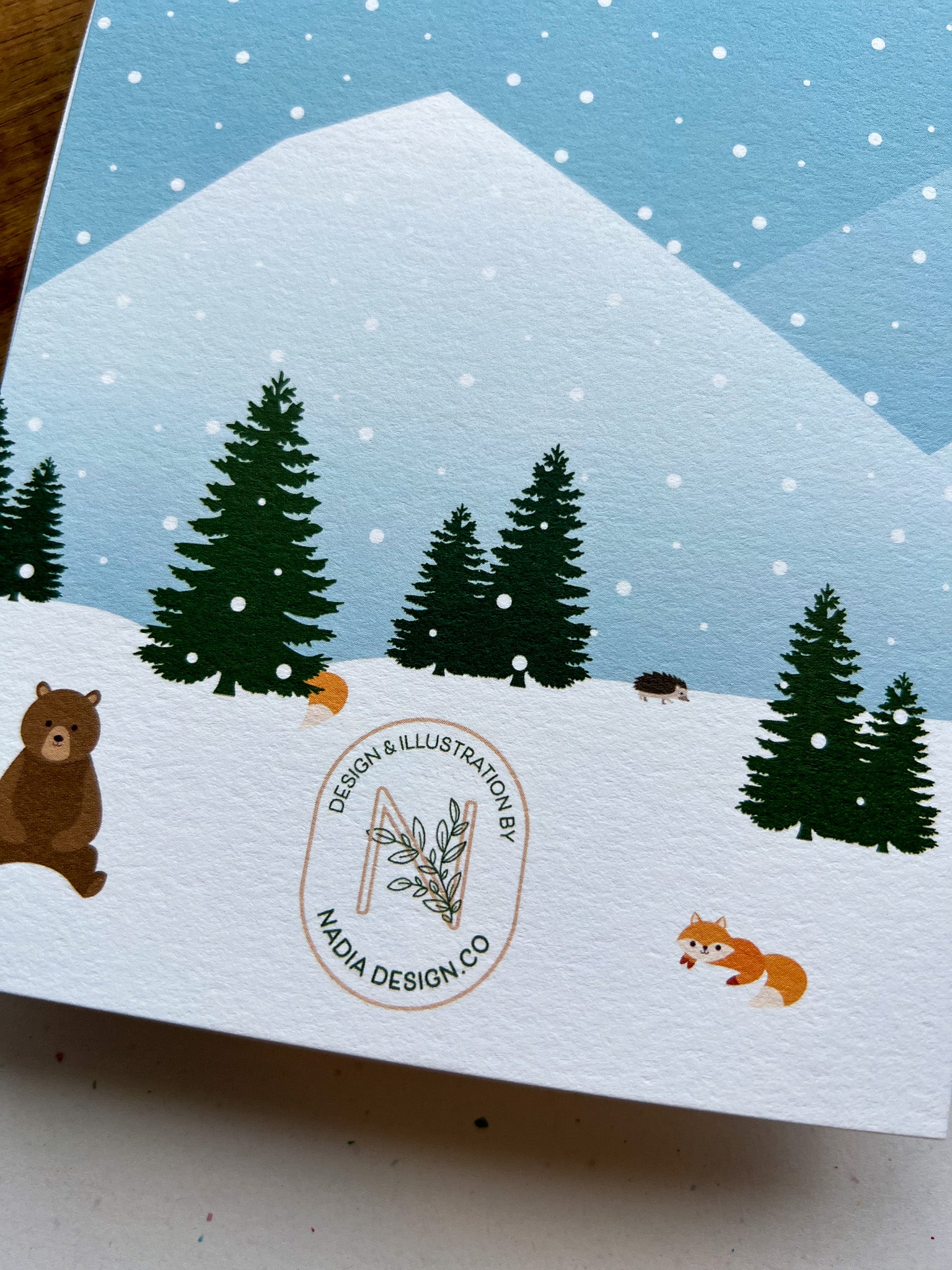 A greeting card depicting a winter scene with a cabin, snow-covered trees, mountains in the background, and a bear in the foreground. The card is placed next to a wooden object, and an envelope with a plastic sleeve is visible.