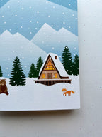A greeting card depicting a winter scene with a cabin, snow-covered trees, mountains in the background, and a bear in the foreground. The card is placed next to a wooden object, and an envelope with a plastic sleeve is visible.