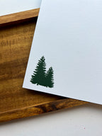 A greeting card depicting a winter scene with a cabin, snow-covered trees, mountains in the background, and a bear in the foreground. The card is placed next to a wooden object, and an envelope with a plastic sleeve is visible.