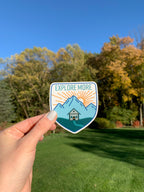 A vinyl sticker with an illustration of a cabin in a mountainous landscape with the text 'EXPLORE MORE' 