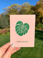 A greeting card with a green leaf illustration and the text 'UNBELEAFABLE' on it, presented with a white envelope and a plastic sleeve.