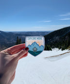A vinyl sticker with an illustration of a cabin in a mountainous landscape with the text 'EXPLORE MORE' held against a blue sky and mountain backdrop.
