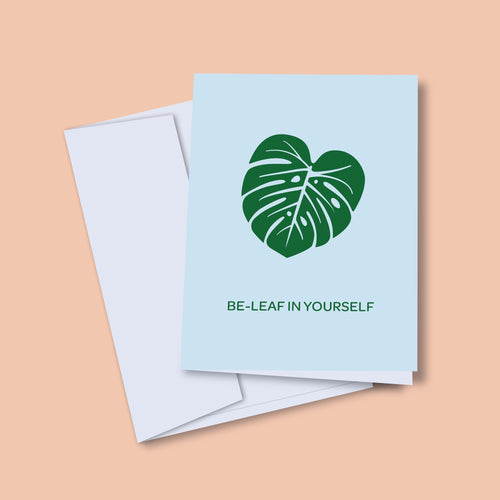 A greeting card with an illustration of a green monstera leaf on it, alongside a white envelope and a plastic sleeve, placed on a wooden surface with some leaves scattered around.