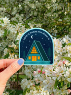 A sticker with an illustration of a cabin under the moon and stars held against a blurred naturxal background.