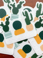 A clear sticker with a pun related to cacti, held against a backdrop of green cacti. The sticker features an illustration of a cactus with the text 'LOOKIN' SHARP'.