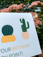 A greeting card with illustrations of three cacti in pots and the text 'HOPE YOUR BIRTHDAY IS ON POINT!' on it, placed on a  with leaves around.