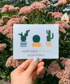 A greeting card with illustrations of three cacti in pots and the text 'HOPE YOUR BIRTHDAY IS ON POINT!' on it, placed on a wooden surface with leaves around.