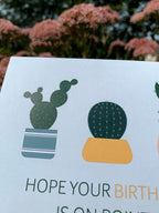 A greeting card with illustrations of three cacti in pots and the text 'HOPE YOUR BIRTHDAY IS ON POINT!' on it, placed on a with leaves around.