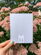 A greeting card with a green and white illustration of a plant, held in a hand with a pink floral background.