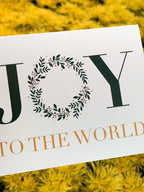 A hand holding a Christmas card with the text 'JOY TO THE WORLD' surrounded by a green leafy illustration, presented against a leafy background.