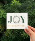A hand holding a Christmas card with the text 'JOY TO THE WORLD' surrounded by a green leafy illustration, presented against a leafy background.