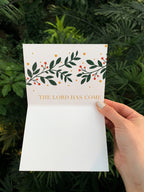 A hand holding a Christmas card with the text 'the lord has come' surrounded by a green leafy illustration, presented against a leafy background.