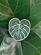 A magnet shaped like a Monstera leaf with the text 'LEAF ME ALONE' in the center, placed on a green leaf.