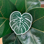 A magnet shaped like a Monstera leaf with the text 'LEAF ME ALONE' in the center, placed on a green leaf.